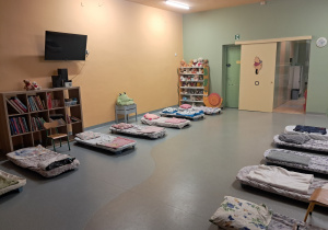 Sala przedszkolna z ustawionymi łóżeczkami, pluszowymi zabawkami i ksiąkami na półkach szafek