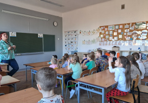 Dzieci w ławkach szkolnych odpowiadają na pytania nauczycielki