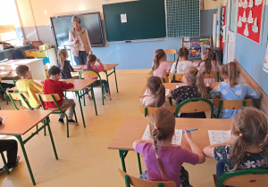 Dzieci siedzą w ławkach szkolnych, nauczycielka spogląda na wykonywane zadanie jednego z dzieci