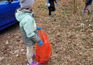 Jedna dziewczynka z workiem na śmieci, a dwie dziewczynki przynoszą jej znalezione w trawie papierki
